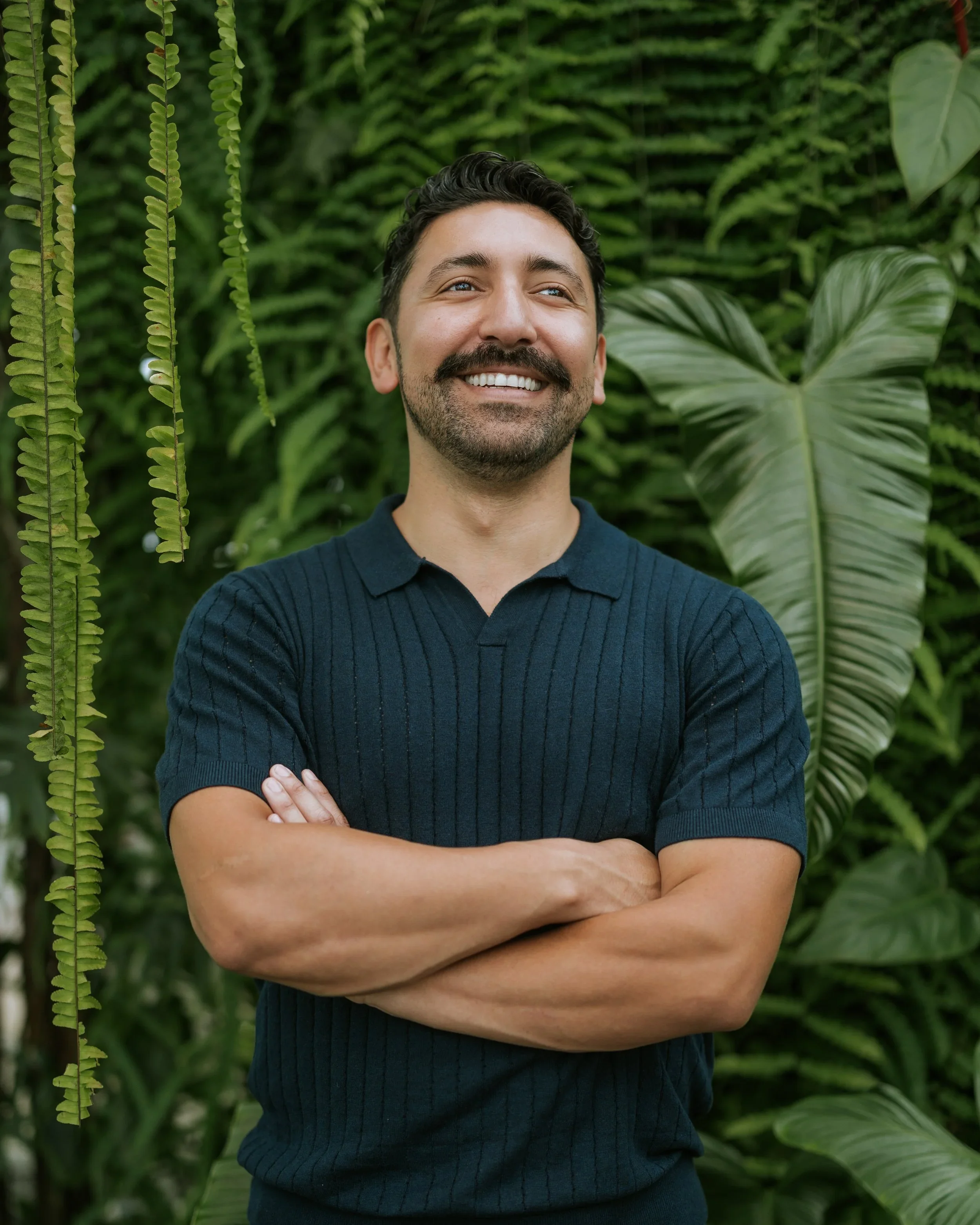 A man with dark hair, a beard, and a mustache, smiling with arms crossed, standing in front of lush green tropical plants.