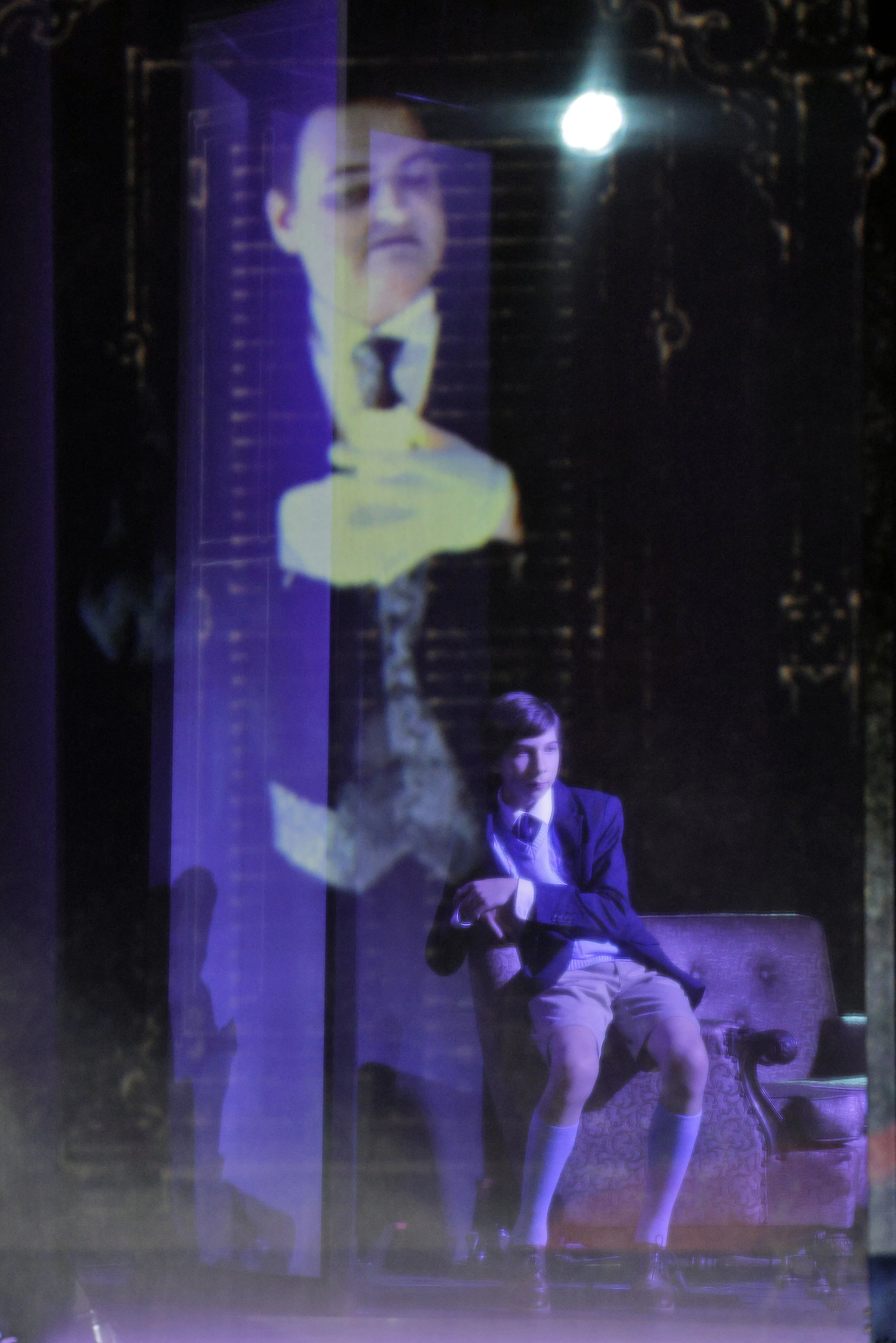 A theatrical scene with two young boys in school uniforms, one sitting on a bench with crossed arms and the other appearing on a screen behind them. The scene has dim lighting and a nostalgic, dramatic atmosphere.