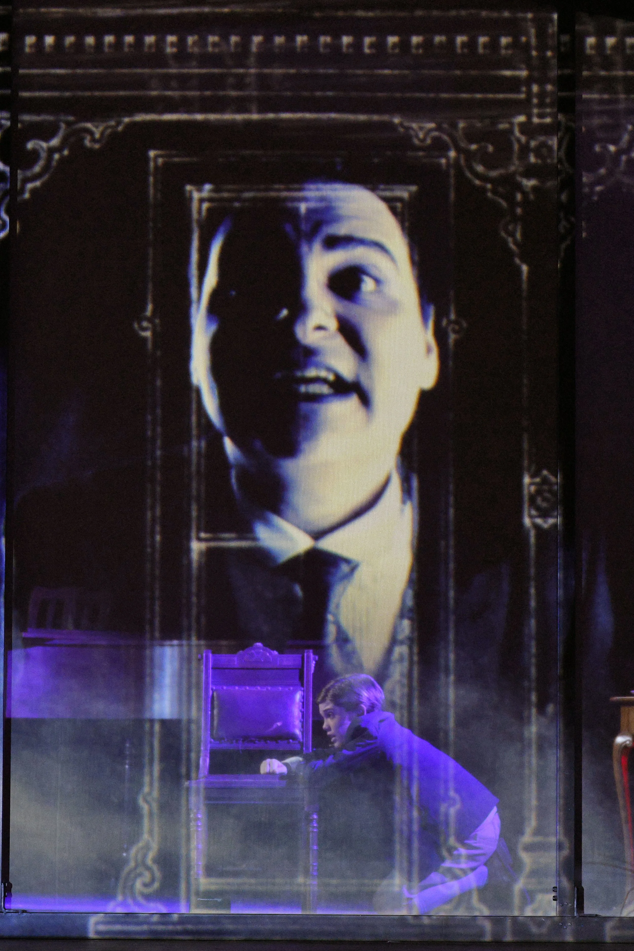 A young boy in a purple jacket kneeling on the floor in front of a stage, looking at a transparent box. Behind him, there is a large poster of a man's face with an expression of surprise or excitement, which is framed with ornate detailing.