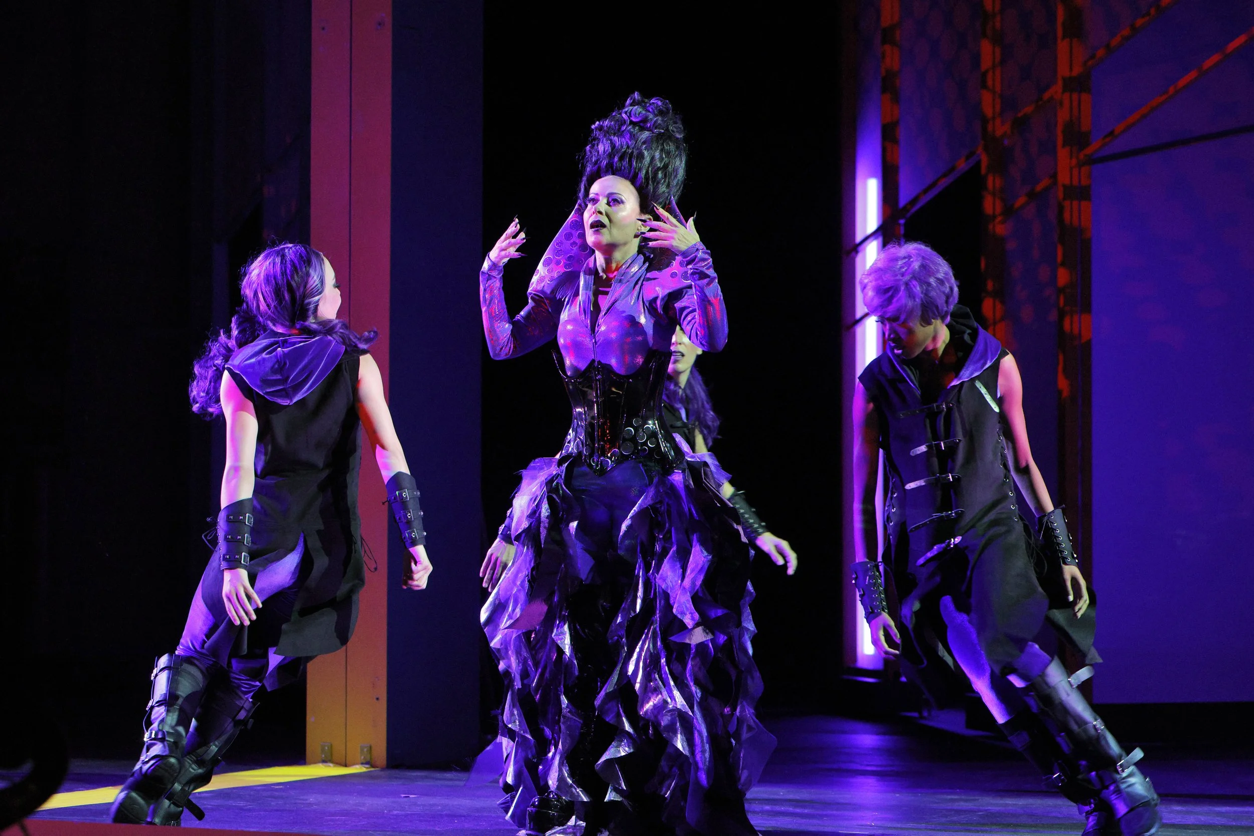 A theatrical performance featuring a woman with dark hair styled in an elaborate updo, dressed in a purple and black costume, flanked by two performers in black outfits with purple accents, on a stage with vibrant lighting and modern set design.