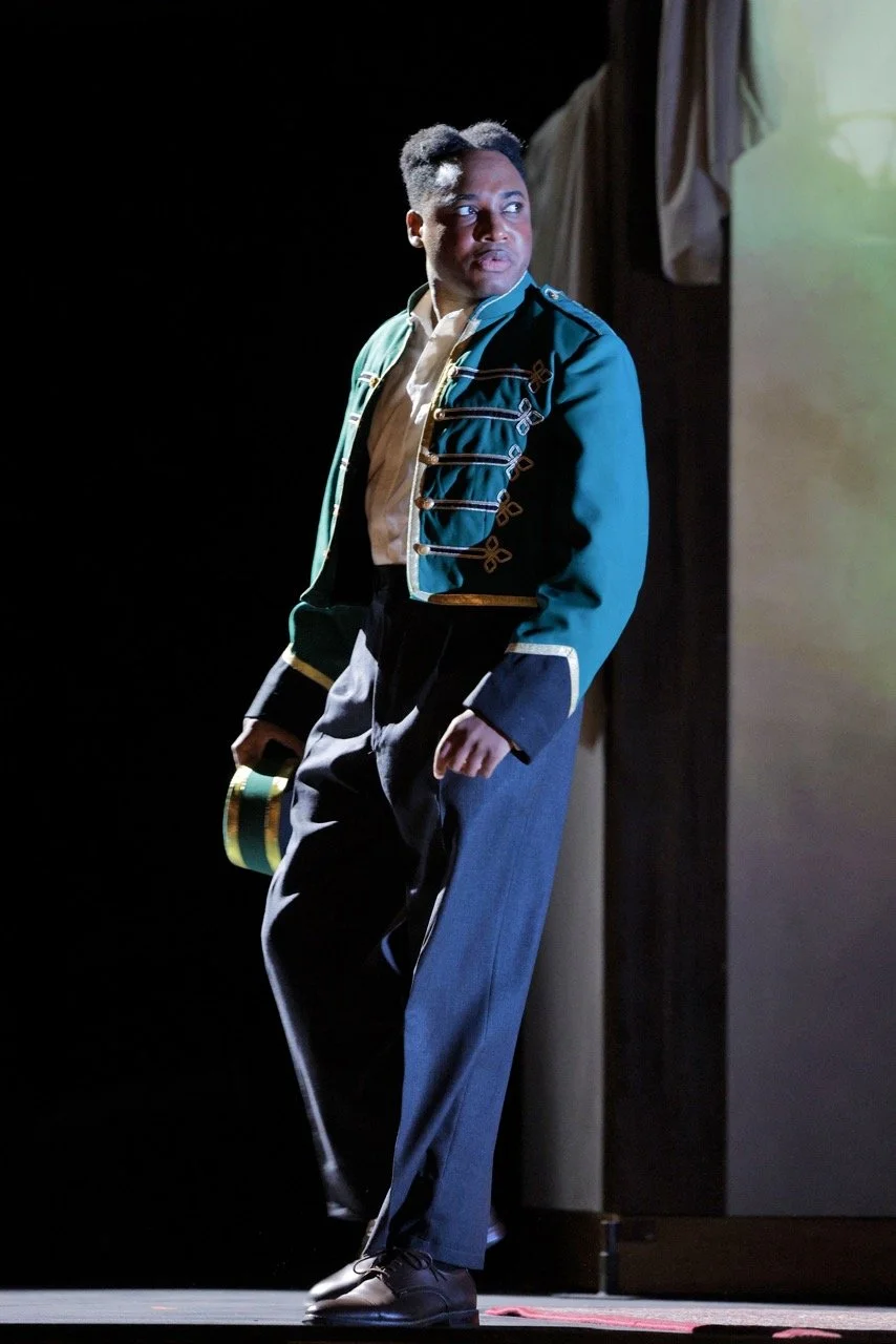 A person dressed in a military-style green jacket with gold trim, beige shirt, dark pants, and black shoes, standing on stage with one hand holding a small green and gold cap, looking to the side with a serious expression.