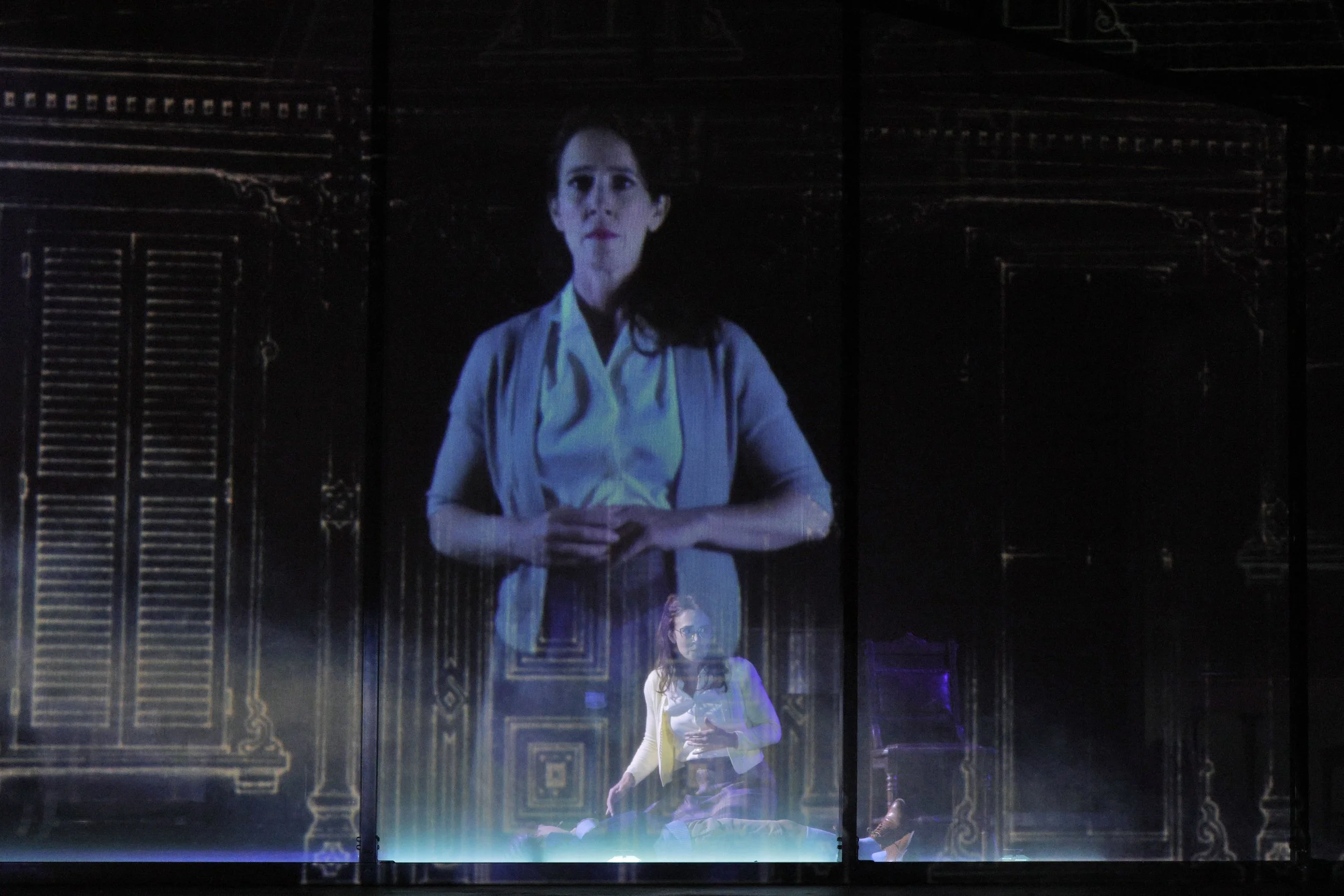 A woman standing in front of a dark ornate background, illuminated with blue lighting, with a semi-transparent reflection or projection of the same woman below her.
