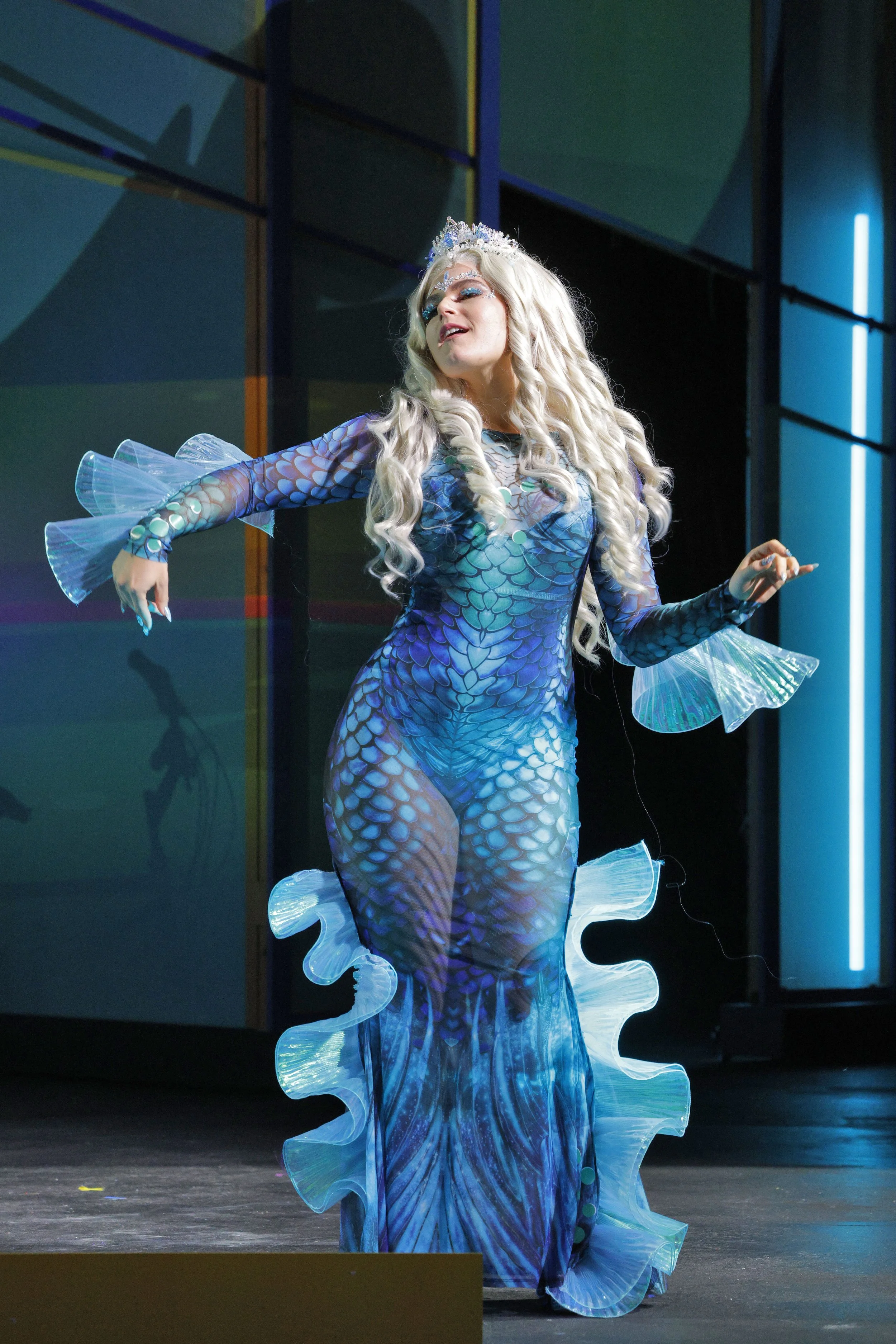 A woman with long, wavy platinum blonde hair performing on stage. She is wearing a blue, iridescent, mermaid-themed dress with ruffled, glowing edges. She is also wearing a tiara and makeup that includes blue eyeshadow and long false eyelashes. The s