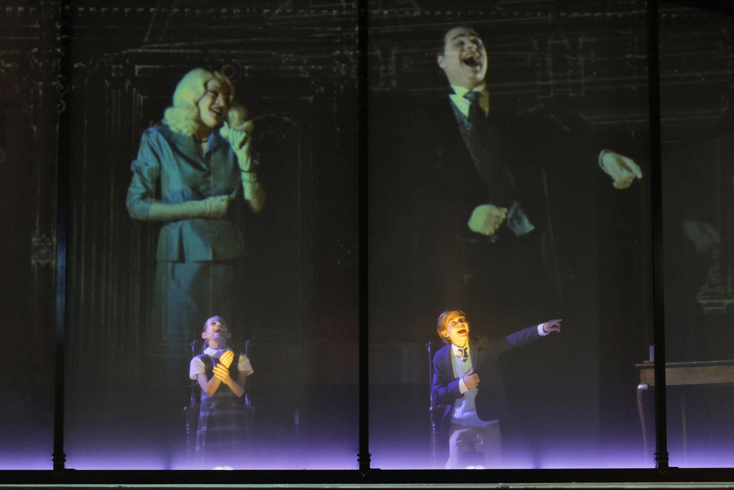 The image depicts a projection of four actors performing on stage. In the projection, a woman with blonde hair in a blue blazer is speaking into a headset microphone, while a man with dark hair and a mustache, wearing a black tuxedo with a bowtie, is