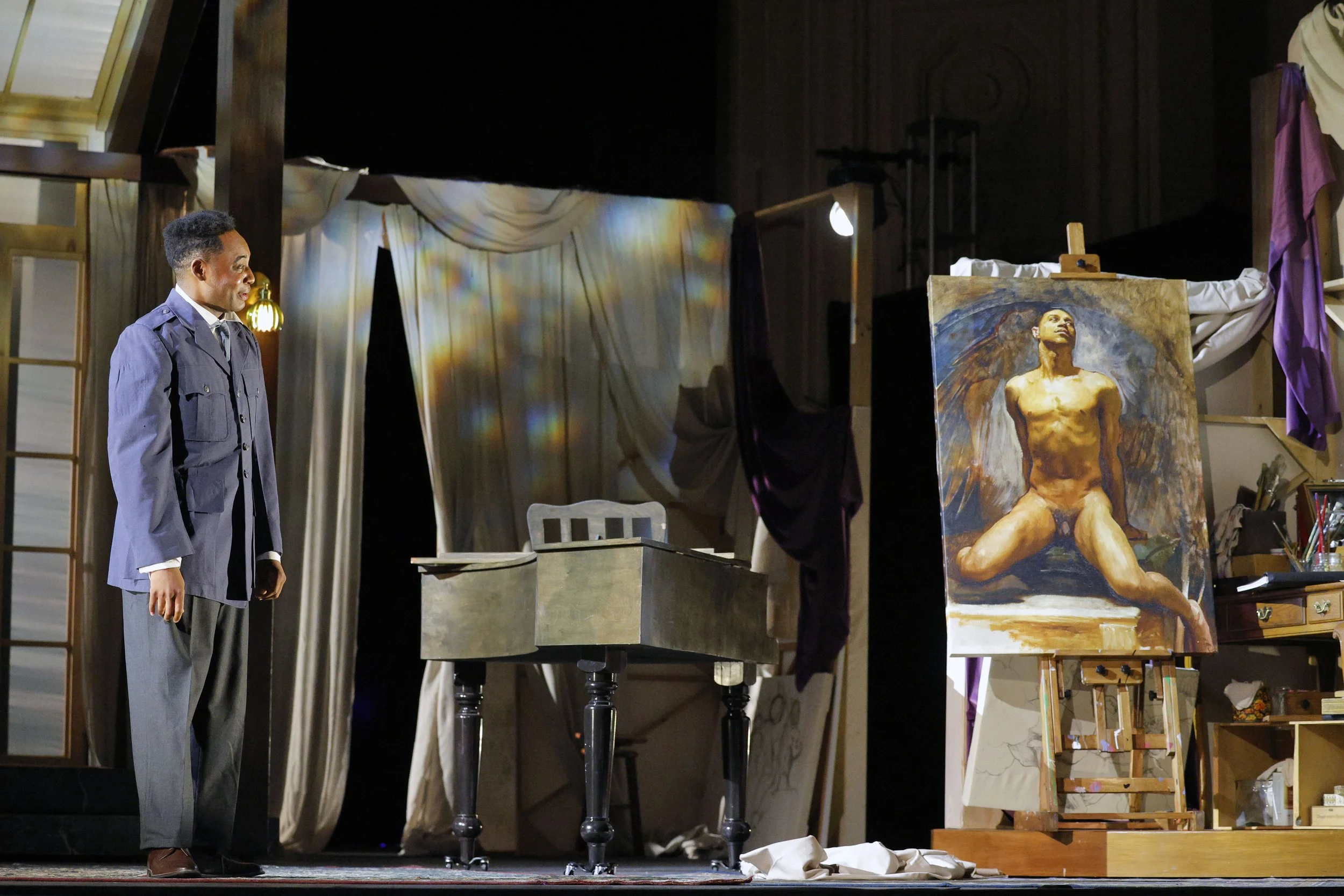 A man in a gray suit standing on a stage next to an easel with a painting of a nude woman in a kneeling pose, with dark hair and a solemn expression, depicted in an expressive, impressionistic style.