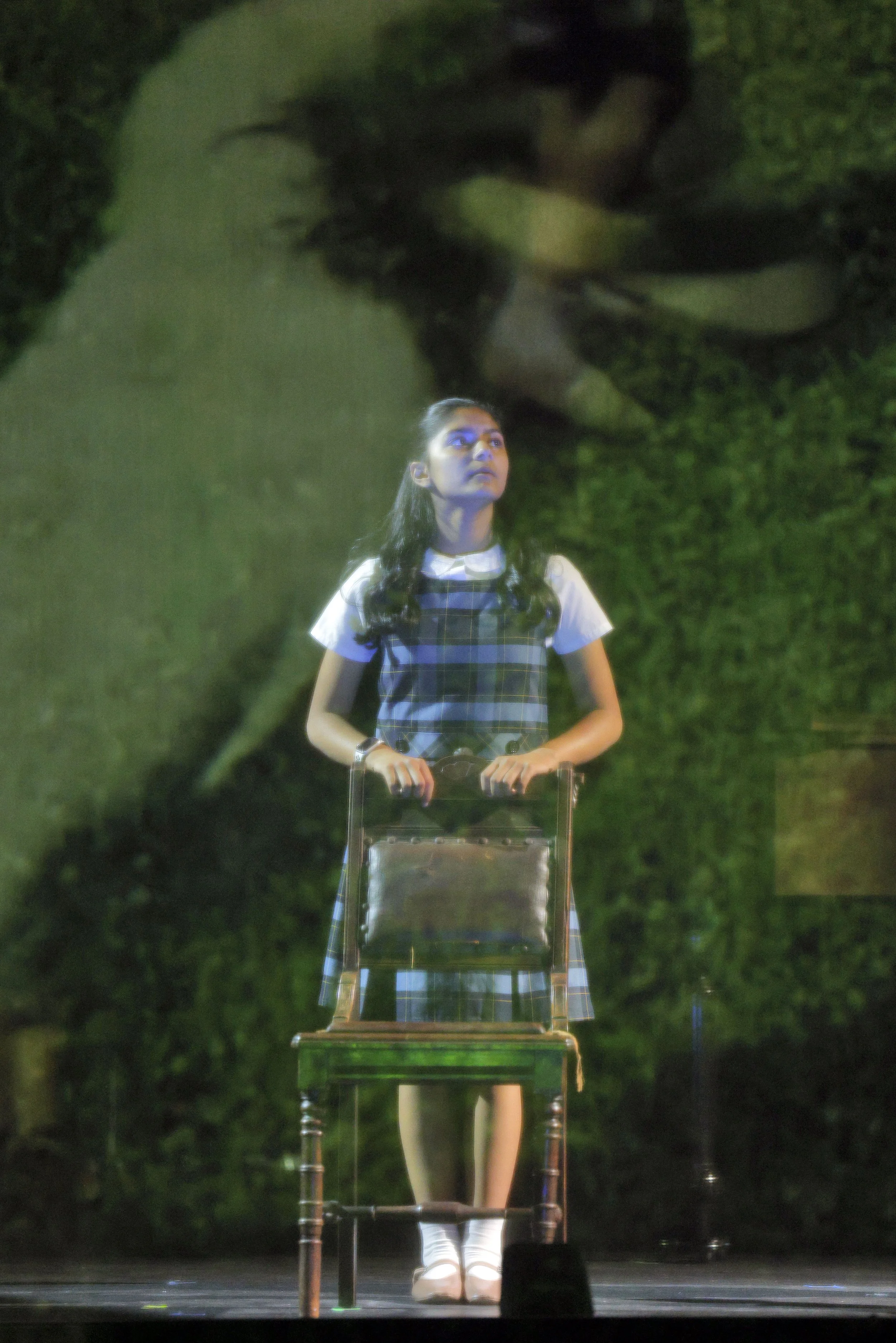 A young woman with long dark hair, wearing a plaid dress over a white shirt, standing on stage in front of a dark green leafy background. She is holding a clear chair with both hands and looking upward with a serious expression.