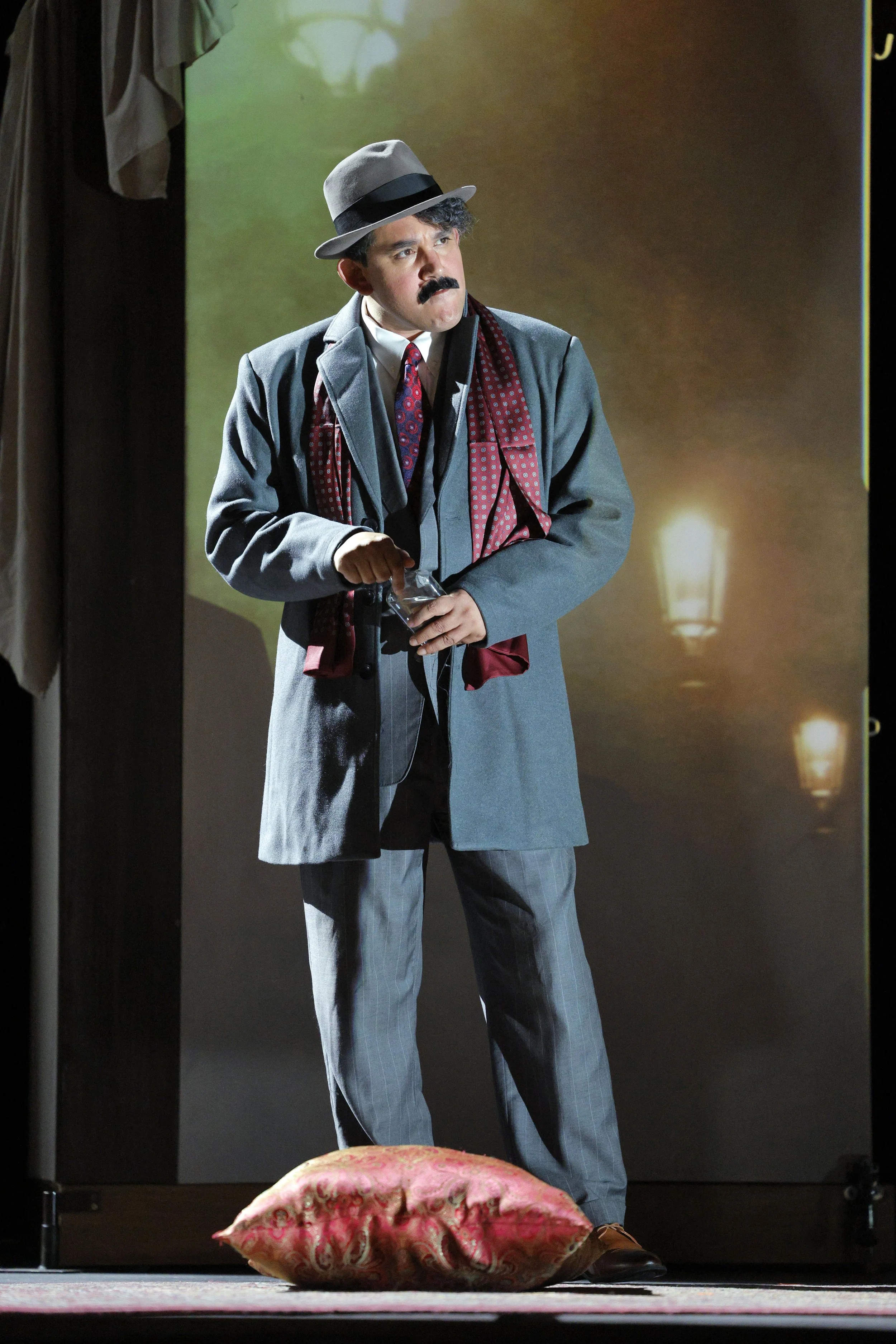 A man with a mustache wearing a gray suit, patterned tie, and a fedora, standing on a stage with a green background. He is holding an object in his hands and looking to the side. There is a decorative pillow on the floor in front of him.