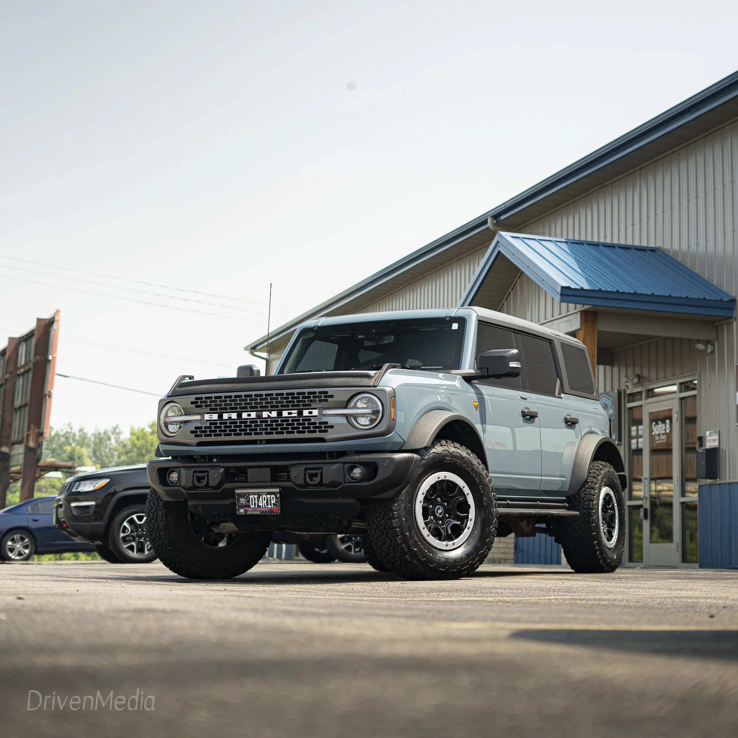 Light blue Ford Bronco parked outside a building with a blue roof, alongside other vehicles, under a clear sky.