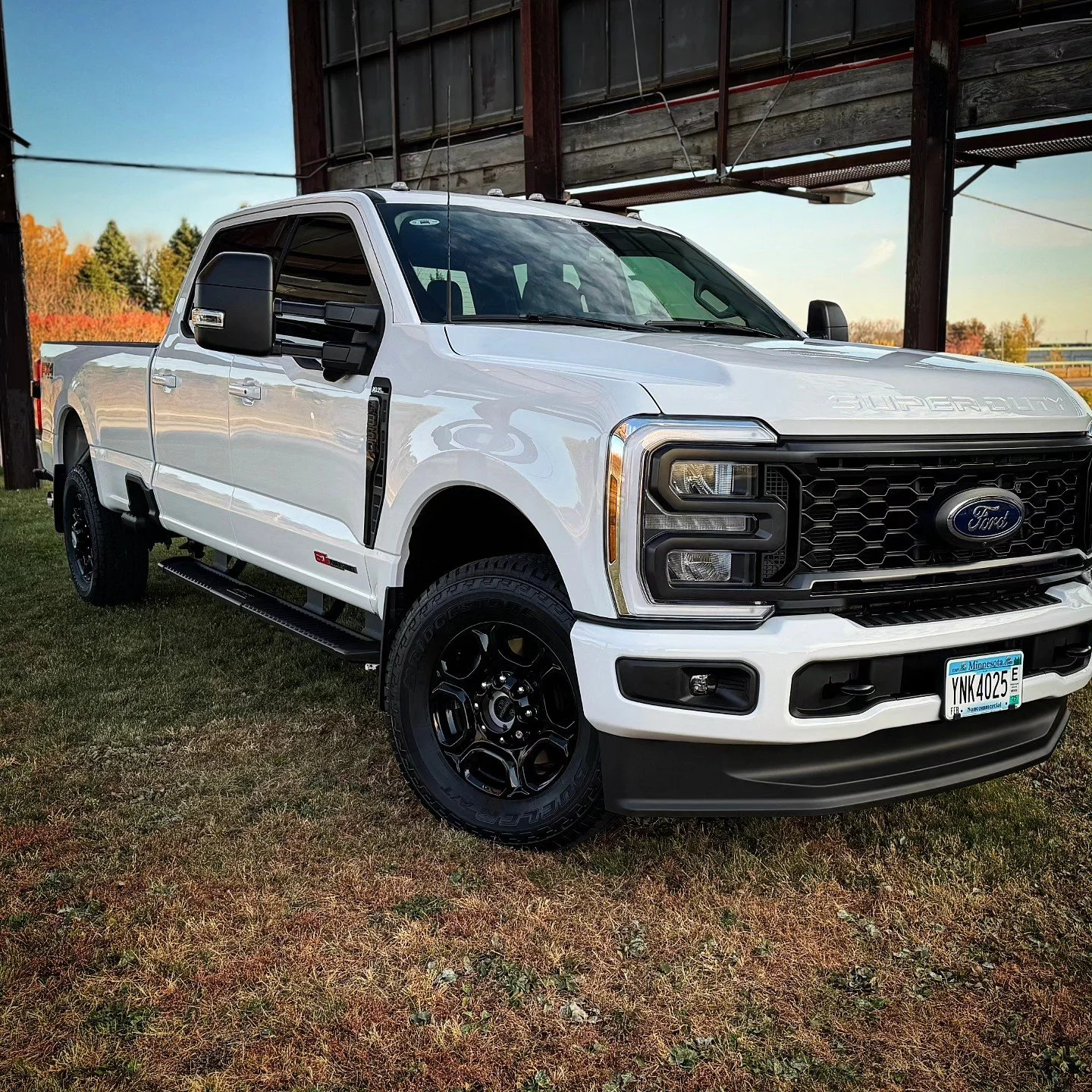 White Ford Super Duty pickup truck parked on grass near a barn or structure with a clear blue sky in the background.