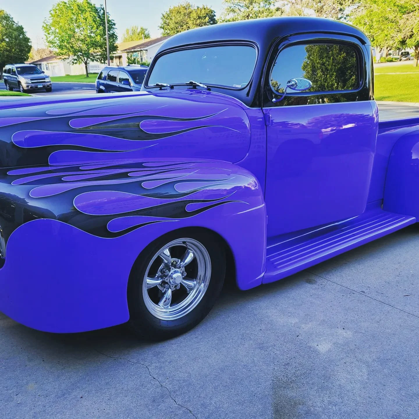 A vintage pickup truck painted in a vibrant purple color with black flame decals on the hood and front fenders, parked on a driveway with other vehicles and green trees in the background.