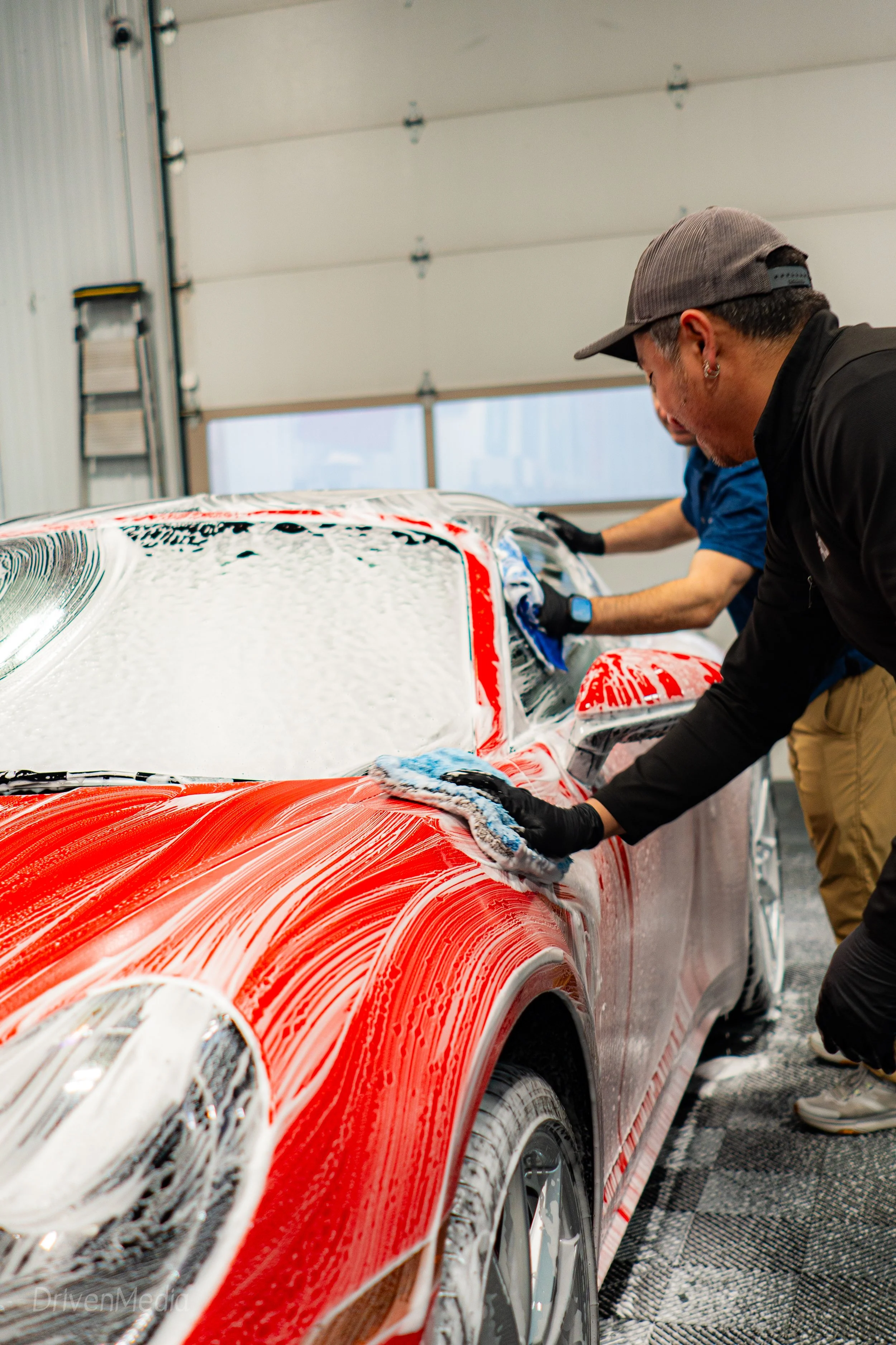 Two people washing a red sports car with soap and sponges inside a garage.