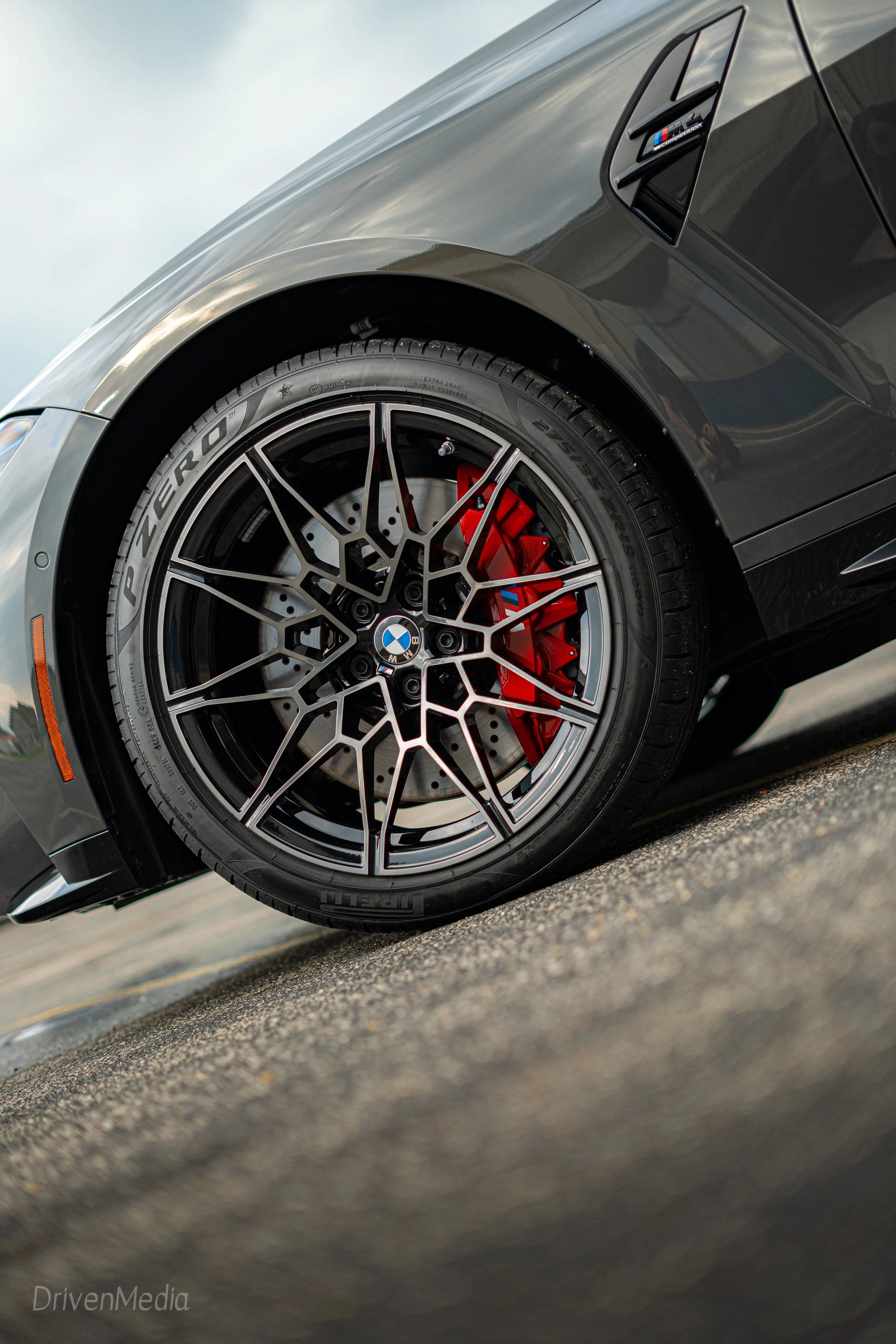 Close-up of a black sports car wheel with a geometric alloy rim, red brake caliper, and BMW logo at the center, on a paved road.