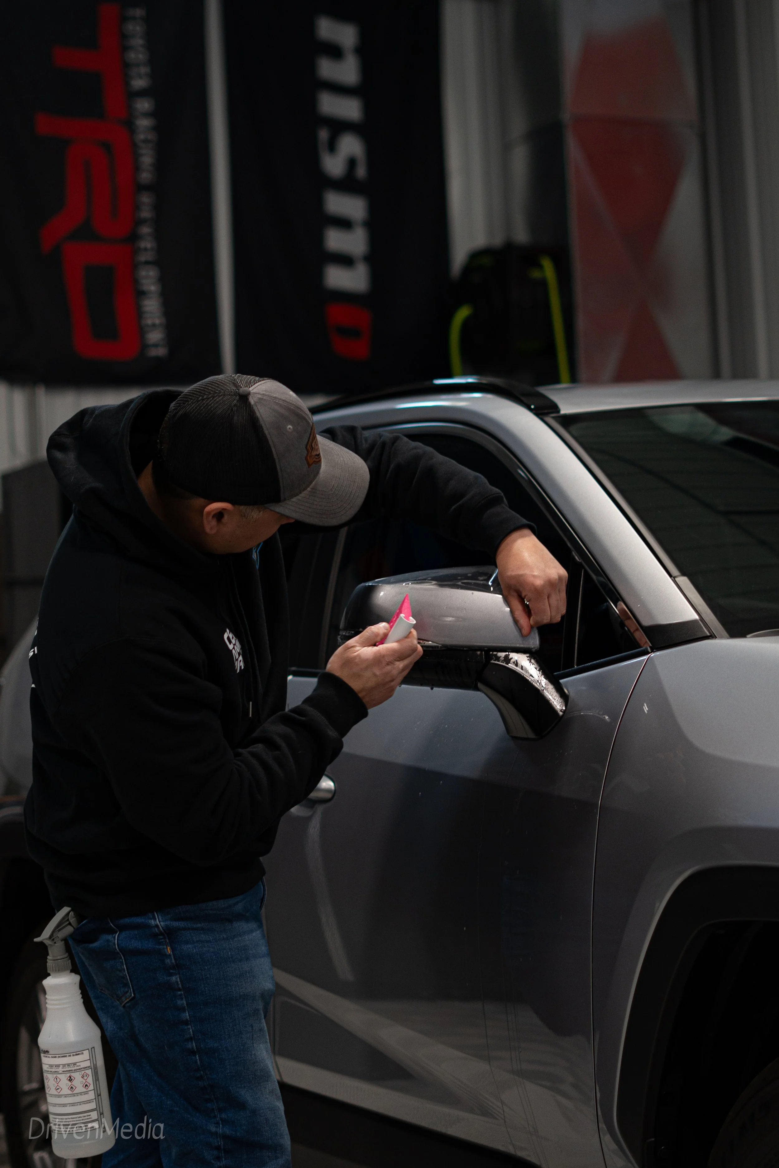 A man applying a protective film to the side mirror of a gray car inside a garage or workshop.