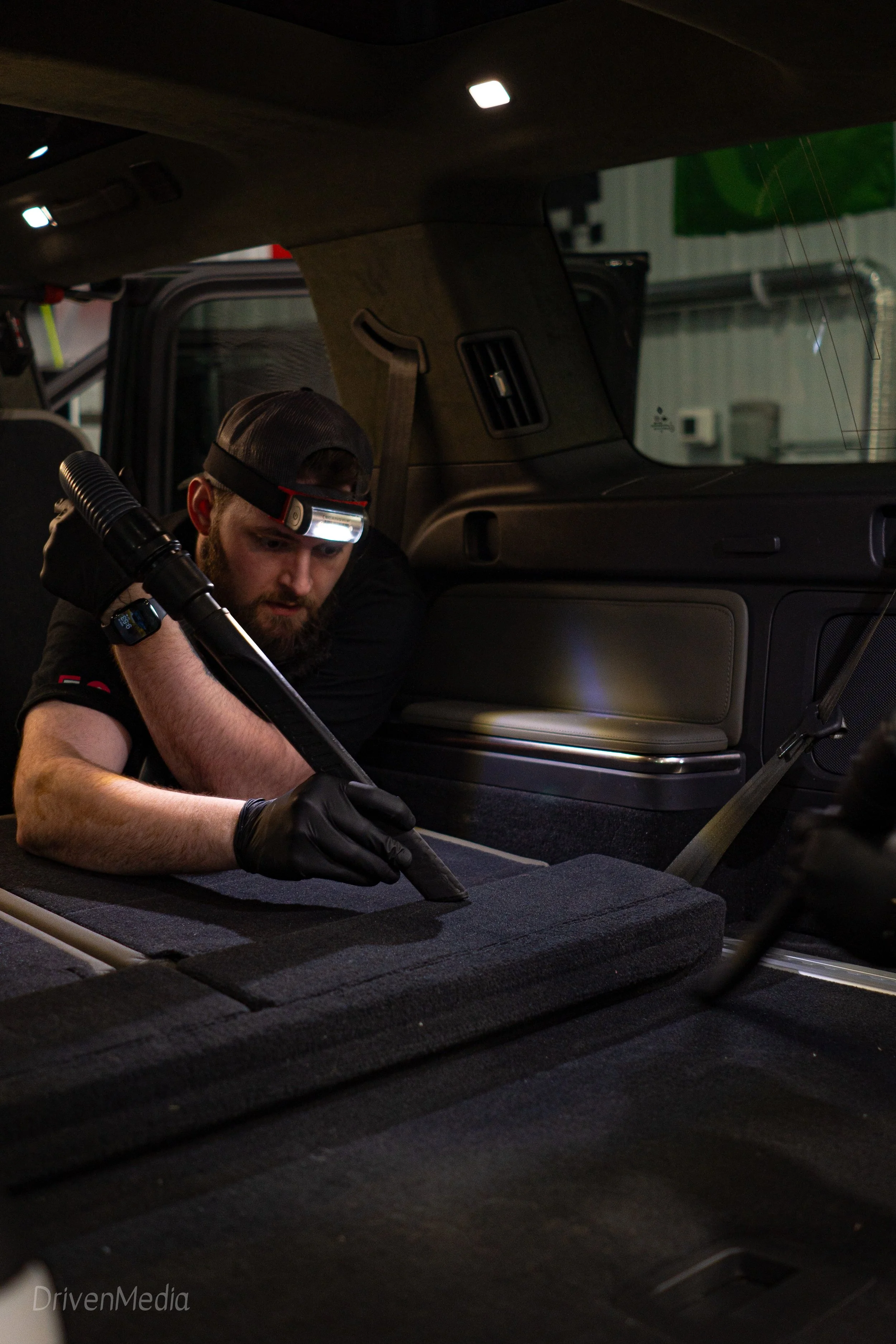A man wearing a headlamp and black gloves is cleaning or inspecting the interior of a vehicle, possibly an SUV, in a garage or workshop.
