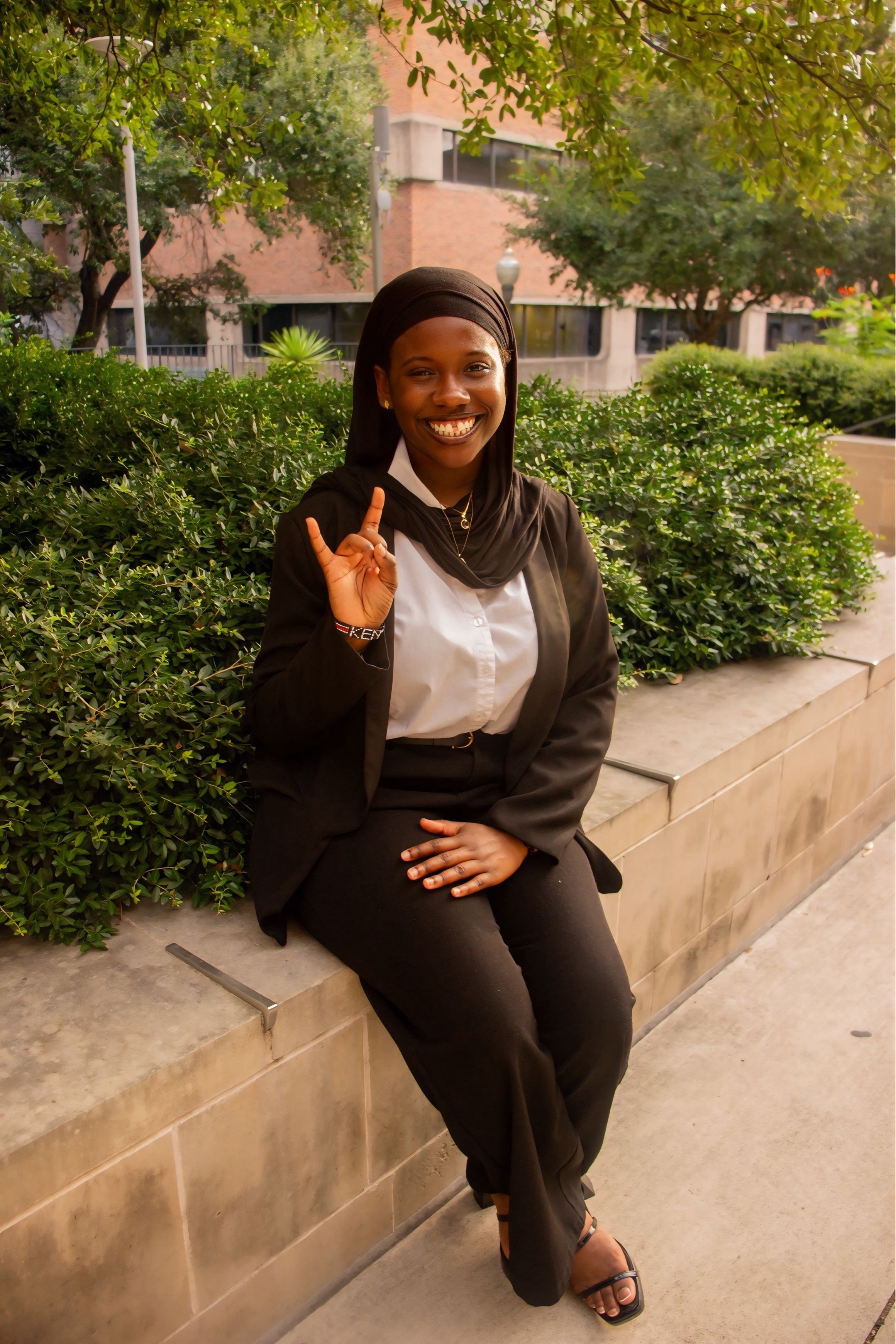 A smiling woman with braided hair, wearing a brown shirt and white vest, standing indoors.