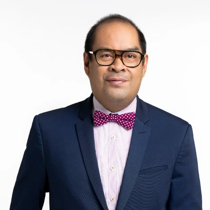 Professional headshot of a man wearing glasses, a navy suit, white shirt, and pink bow tie against a light background.