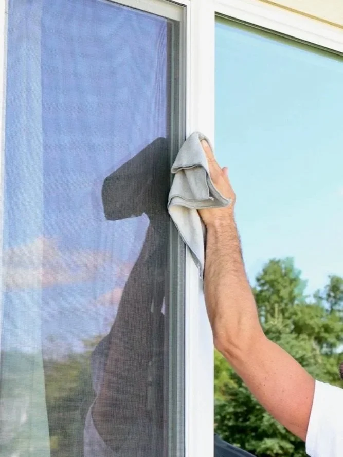 Person cleaning a window with a cloth on a sunny day, outdoors with trees visible in the background.