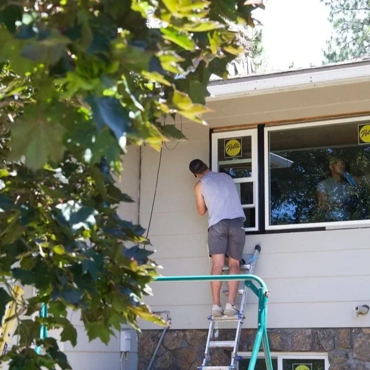 A man installing an exterior glass window from the outside of a house while standing on a ladder, with a reflection of himself in the window.