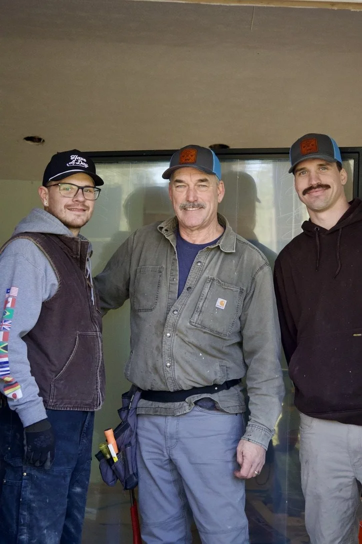 Three men standing side by side indoors, wearing casual work clothes and hats, smiling at the camera.