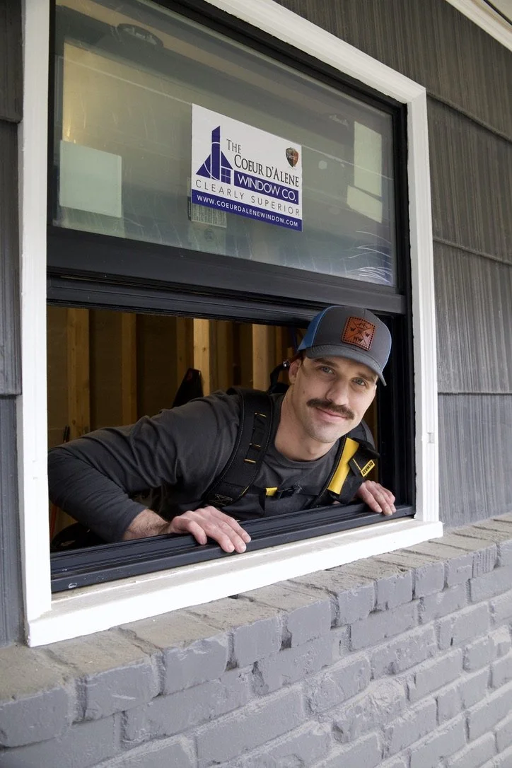 A man with a mustache leaning out of a window, wearing a gray cap and a black shirt, smiling at the camera. The window has a sign for a window company and is part of a gray house with a brick ledge.