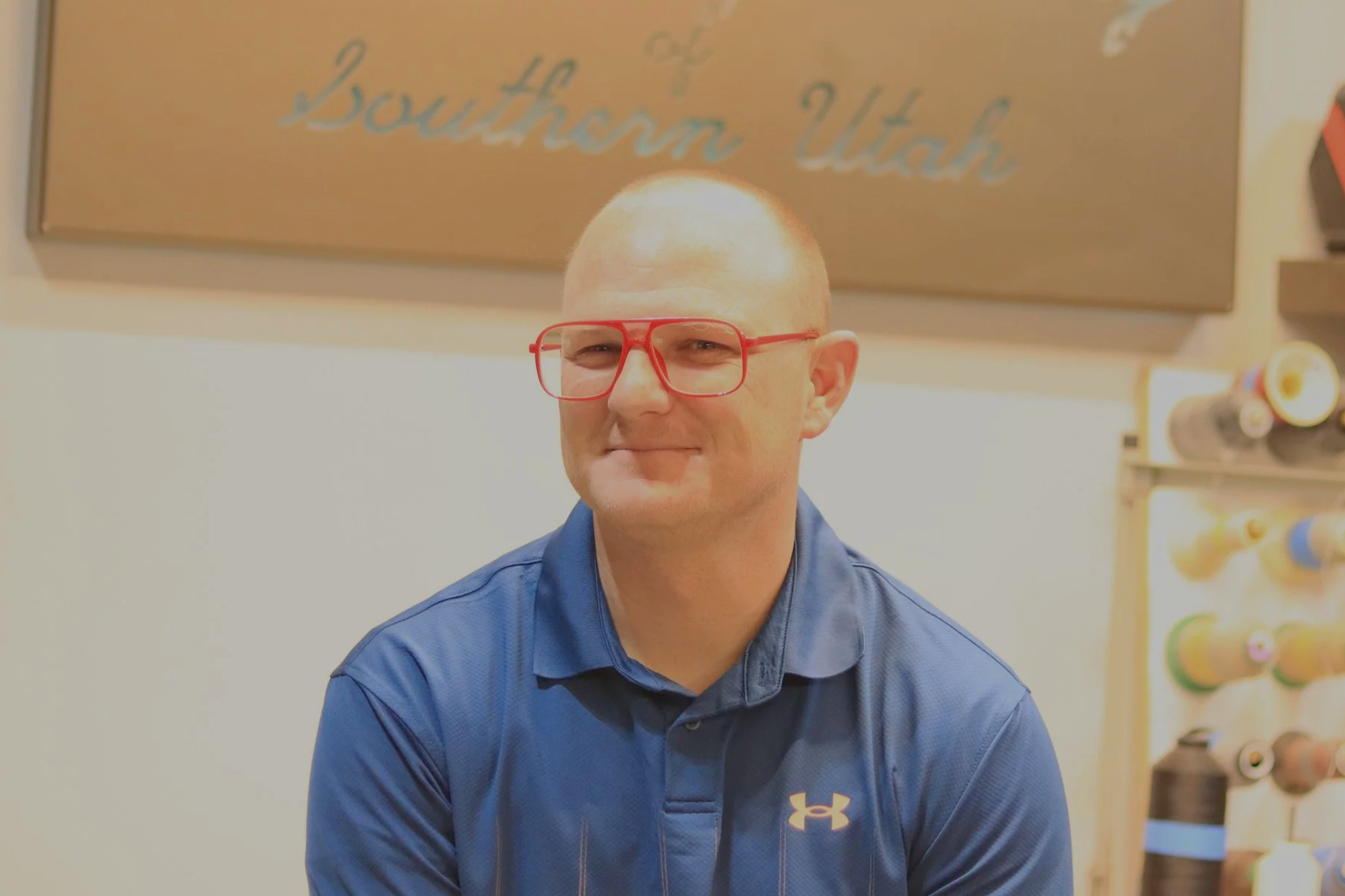 A man with a shaved head wearing red glasses and a blue Under Armour polo shirt, smiling slightly in front of a sign that reads 'Southern Utah'.