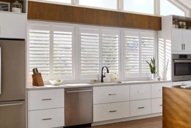 Modern kitchen with white cabinetry, large window with white plantation shutters, stainless steel sink, black faucet, and a potted plant.