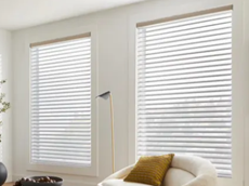 Two large white window blinds partly open on a white wall in a bright room with a beige sofa and a yellow pillow.
