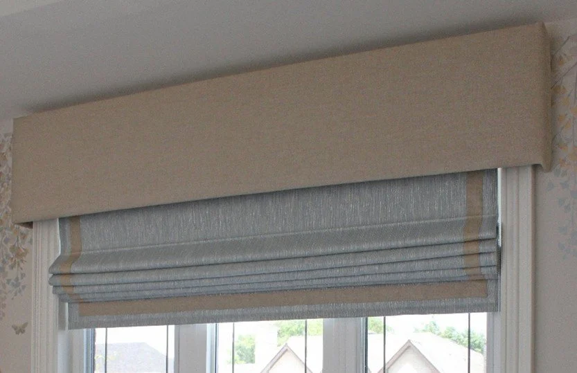 Window with beige valance and grey Roman shades with a beige edge, overlooking houses and trees outside.