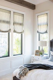 A bedroom with three large windows, a white bedside table with a lamp, and a bed with white and gray bedding.