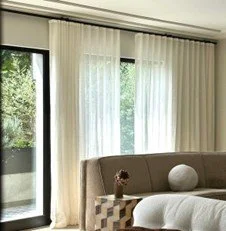 Living room with large windows, white curtains, a beige sofa, a round white decorative object, and a potted plant.