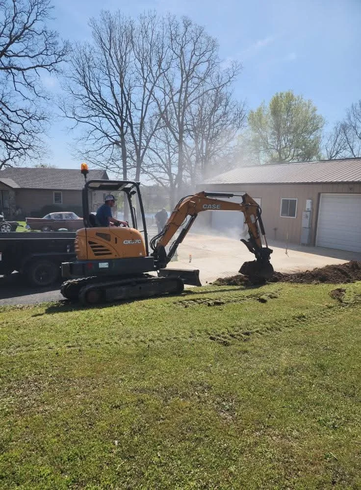 A construction worker operating a small CASE excavator digging up grass and soil in a yard near a driveway and garage, with houses and leafless trees in the background on a clear, sunny day.