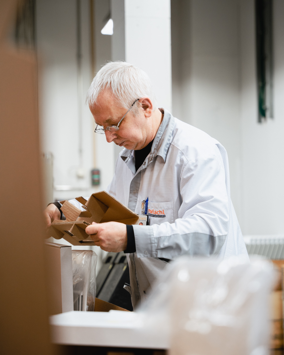 An older man with gray hair and glasses wearing a white shirt looking at a cardboard box in a warehouse or storage area.