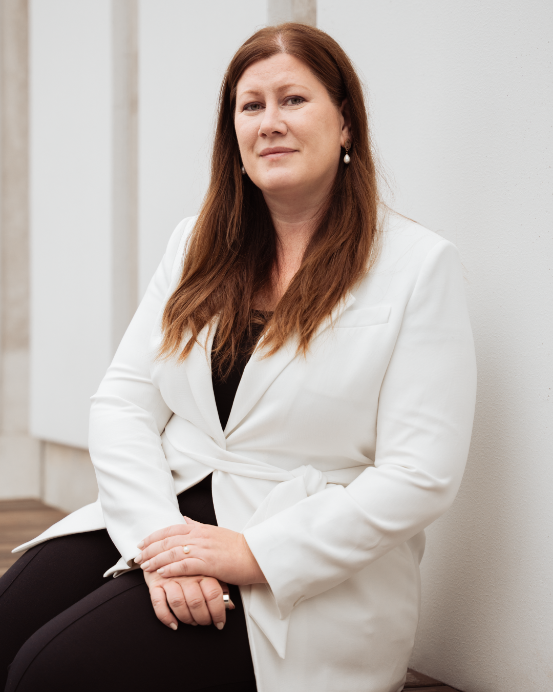 A woman with long red hair sitting against a light-colored wall, wearing a white blazer and black top.