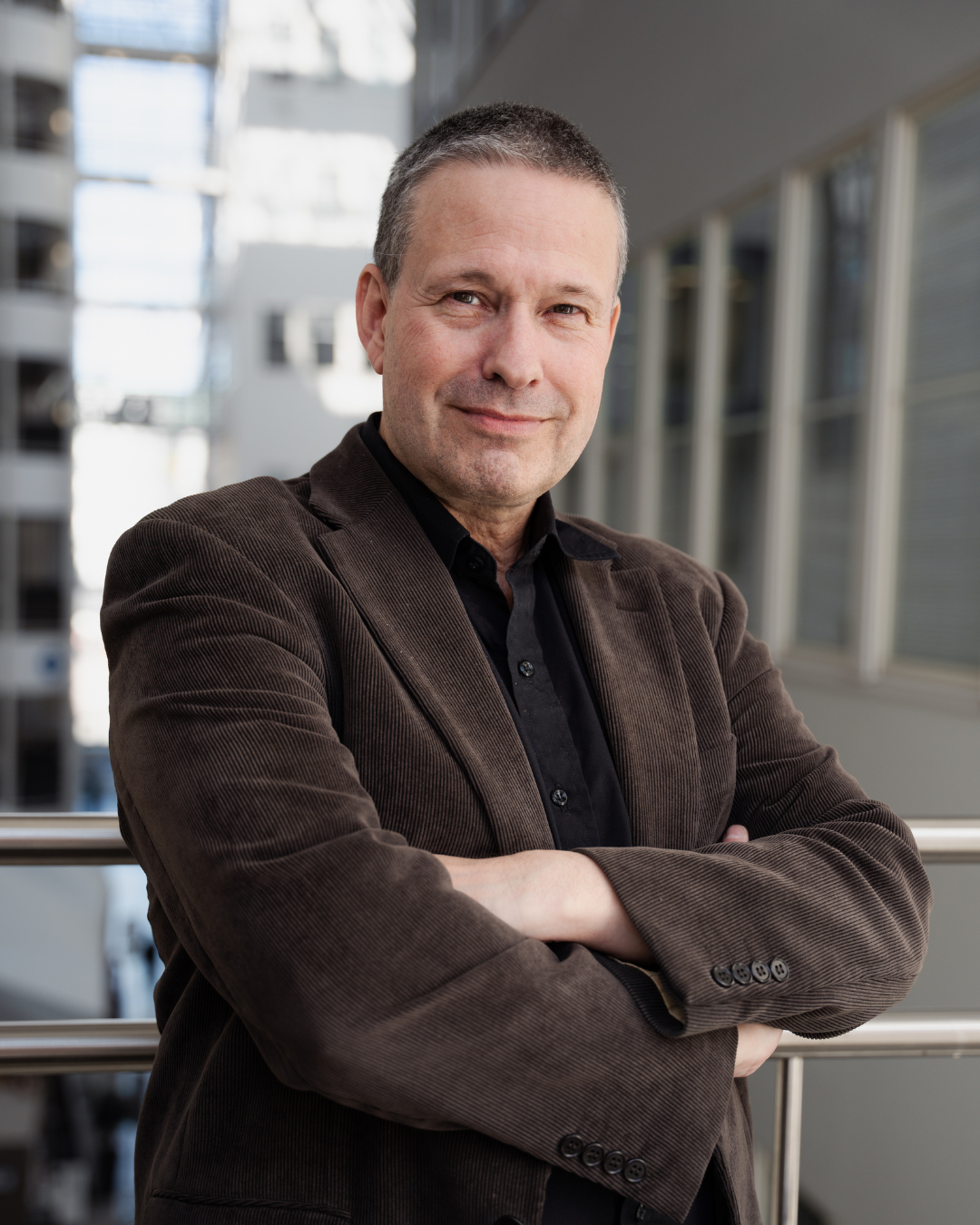 A confident middle-aged man with short gray hair, wearing a dark brown blazer over a black shirt, standing outside by a railing with arms crossed, in an urban setting with modern buildings in the background.