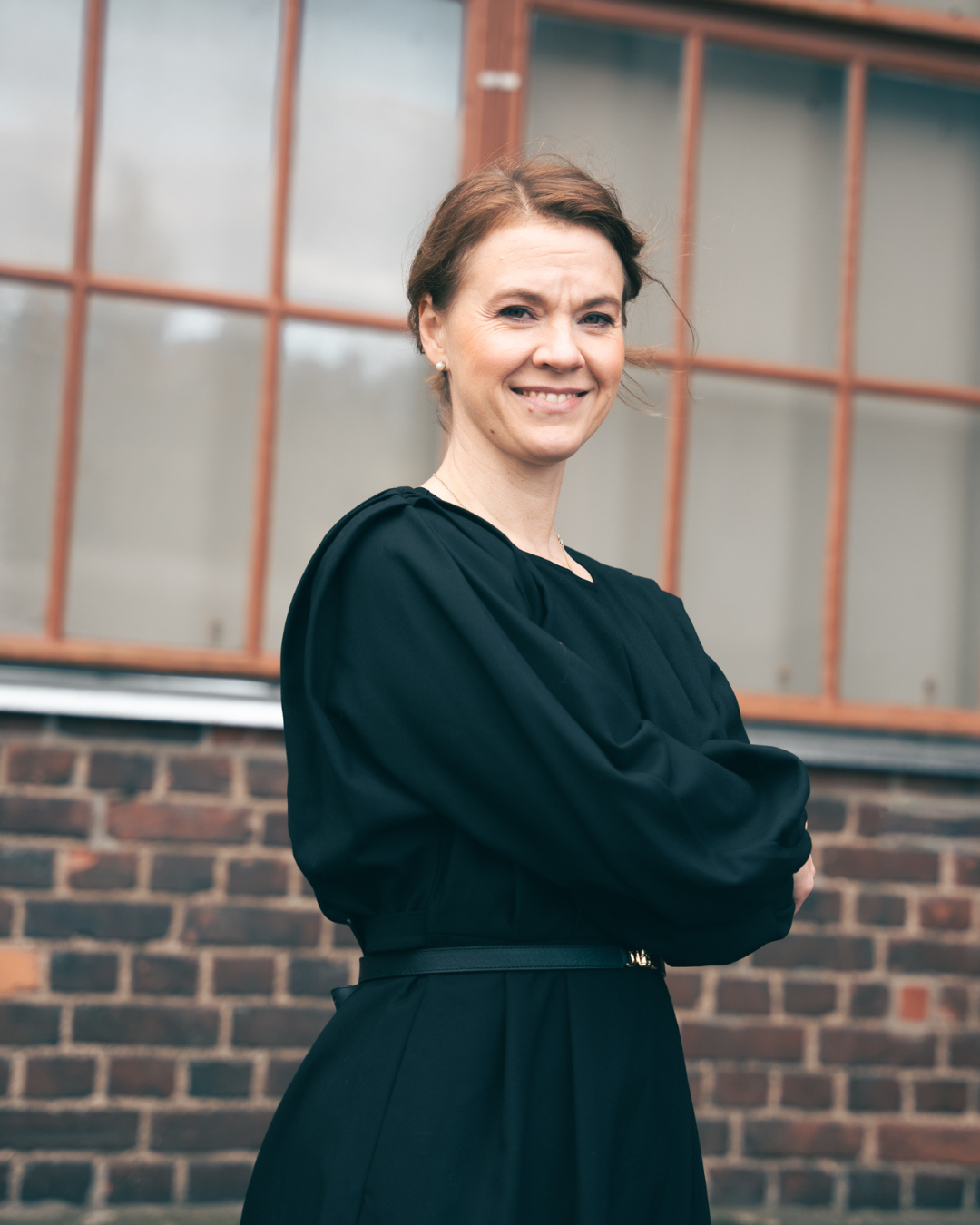 A woman with short brown hair wearing a black dress, standing outside in front of a brick building with large windows, smiling with arms crossed.