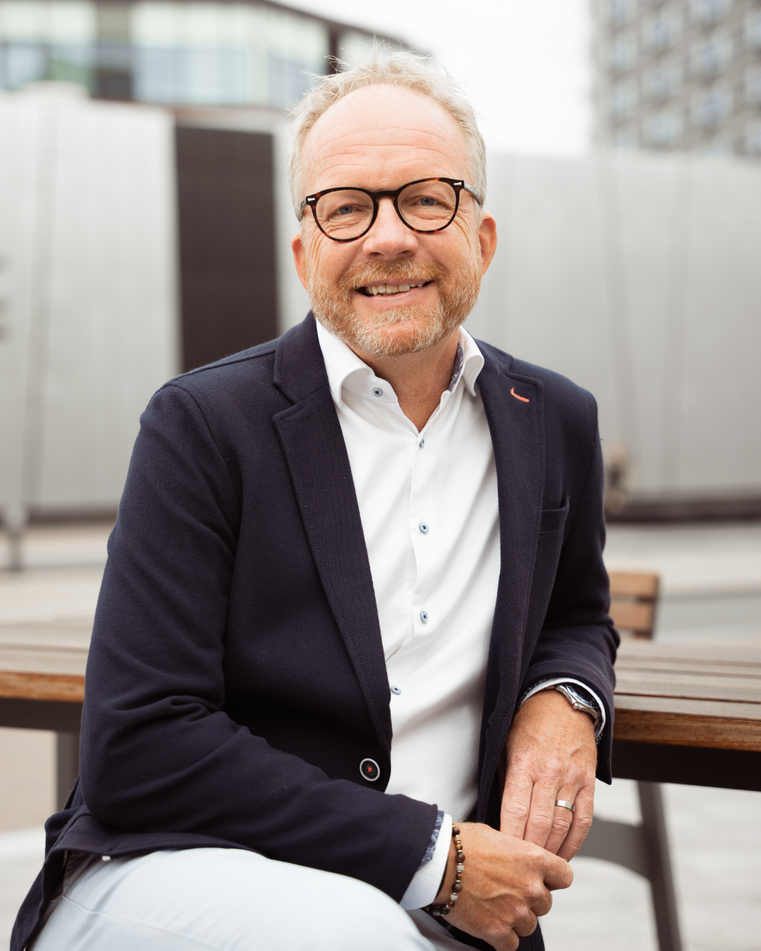 A smiling middle-aged man with glasses, a beard, and light hair, wearing a navy blazer and white shirt, sitting outside at a wooden table in an urban setting.
