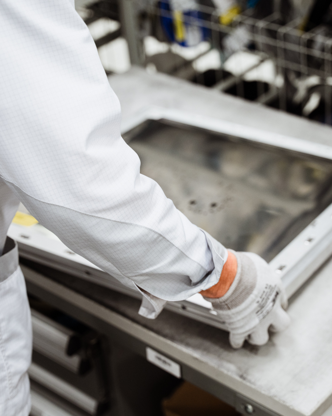 Person working with metal frame or component at a workstation, wearing a white lab coat and gloves.