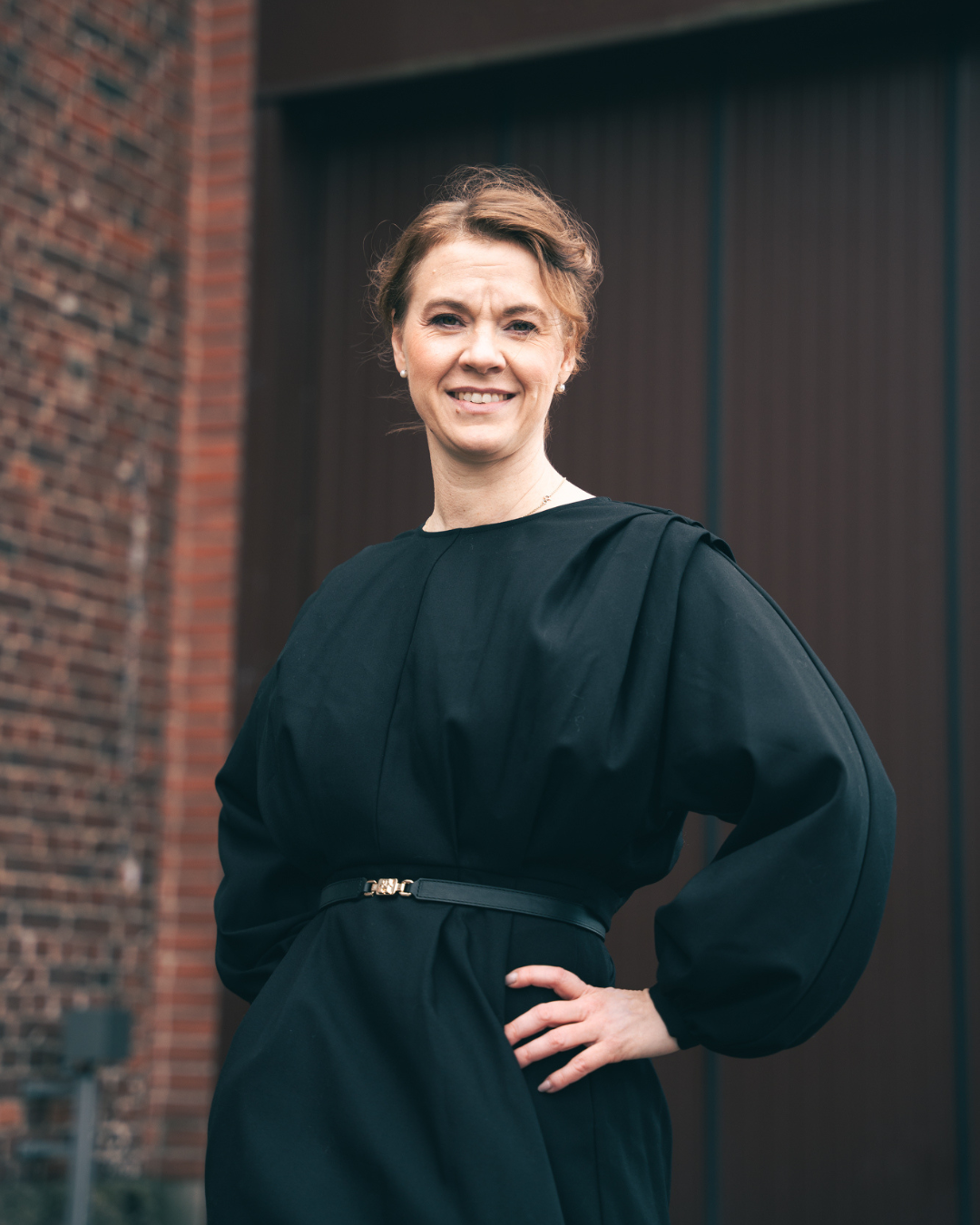 A woman in a black dress standing outdoors with a brick wall and a gate in the background, smiling at the camera.