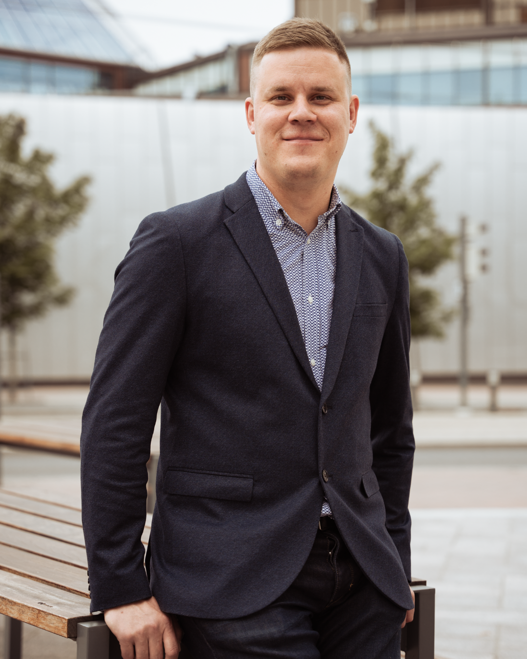 A young man in a business suit posing outdoors with a blurred modern building and trees in the background.