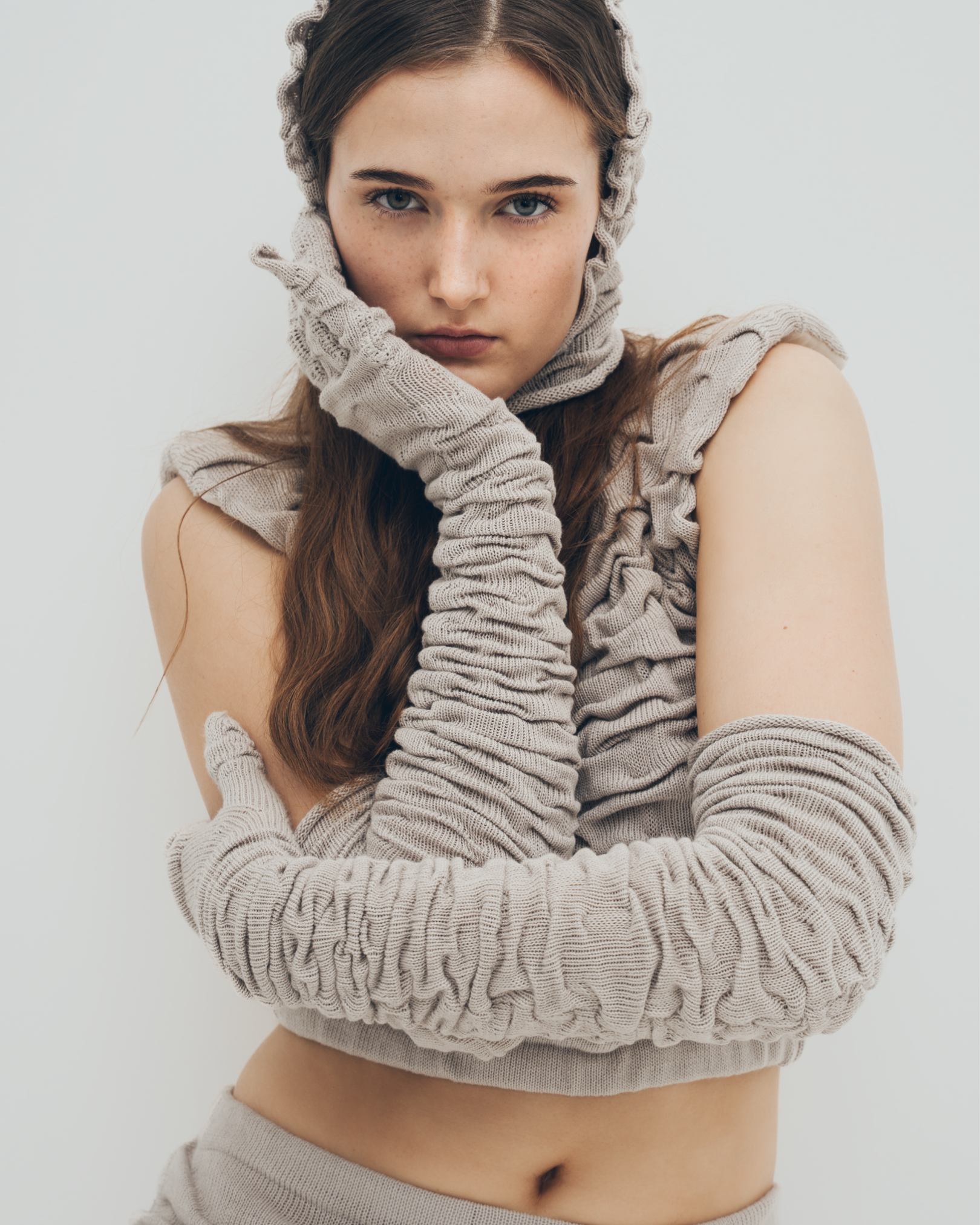 A young woman with long, wavy brown hair and blue eyes wearing a beige knit headscarf, matching gloves, and sleeveless top with ruffled accents, standing against a plain white background.