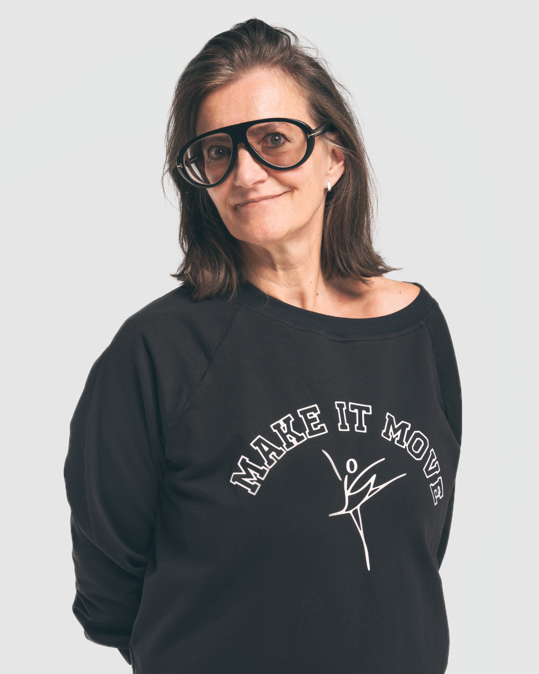 A woman with shoulder-length brown hair wearing large black glasses and a black sweatshirt with the text 'MAKE IT MOVE' and a figure in motion.