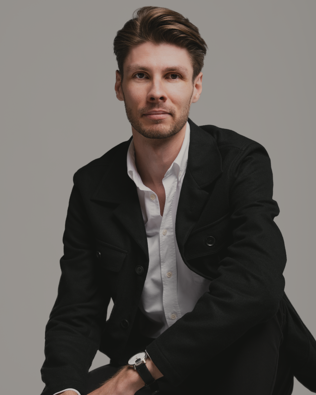 Portrait of a young man with brown hair and a slight beard, wearing a white shirt and black jacket, sitting against a plain gray background.