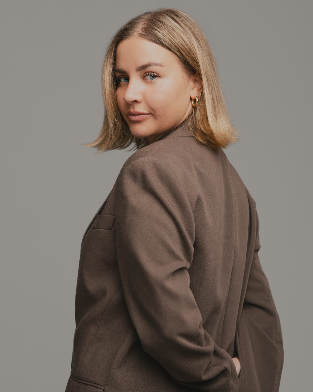 A young woman with shoulder-length blonde hair, blue eyes, and gold hoop earrings, wearing a brown blazer, looking over her shoulder against a gray background.