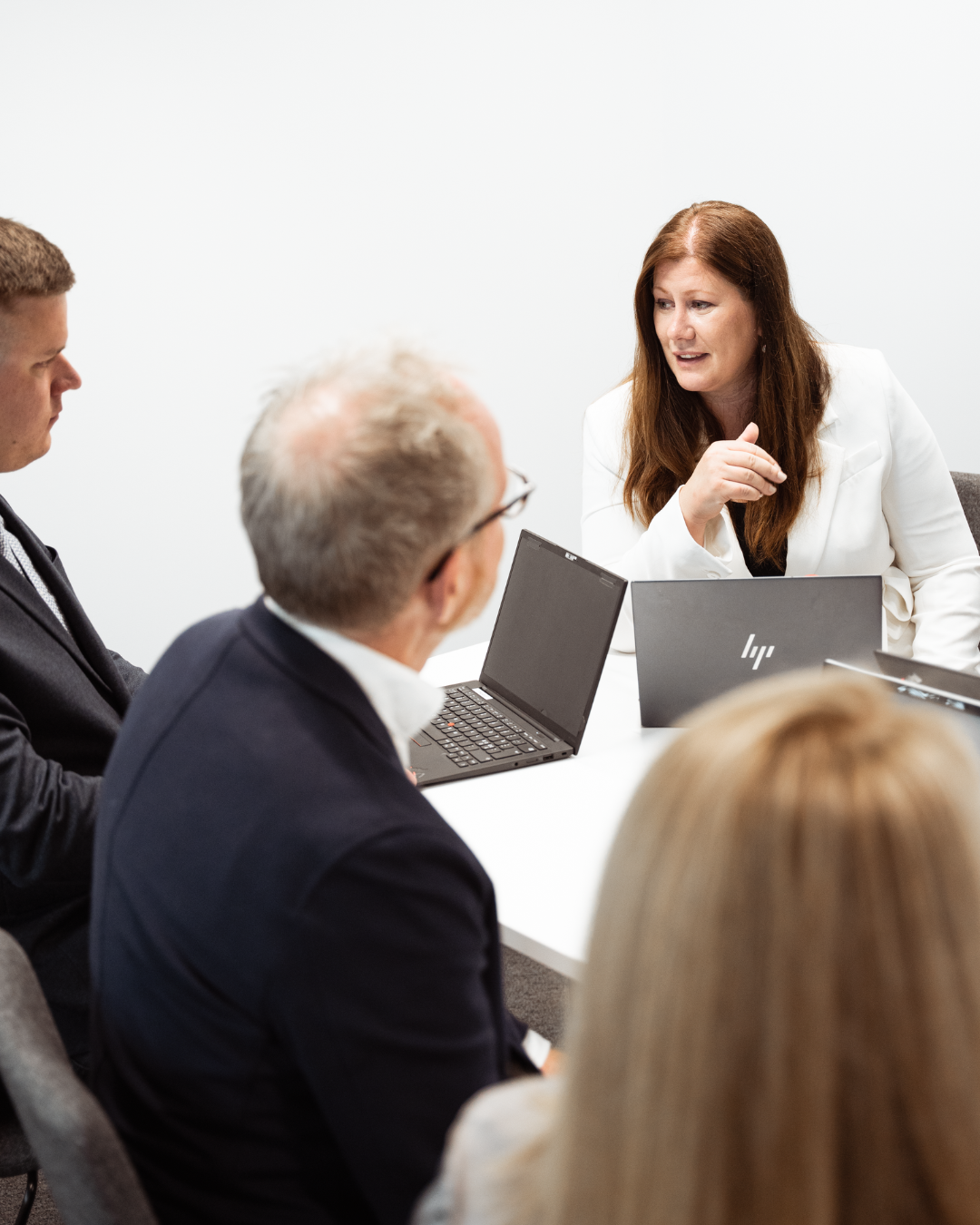 Business meeting with five people, woman speaking, laptops on table, plain white wall background.