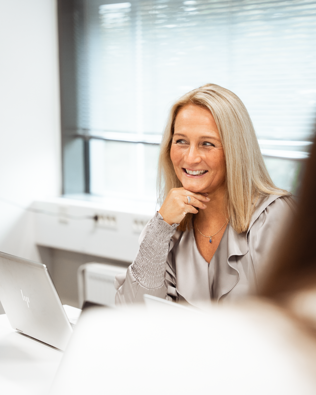 A woman with blonde hair smiling and sitting at a desk with a laptop, in a bright office with window blinds.
