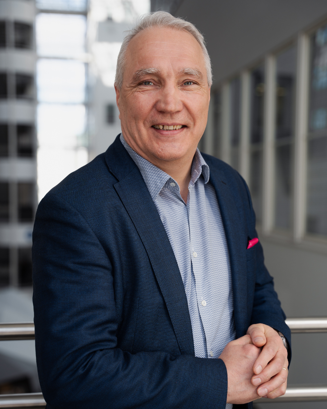A middle-aged man with gray hair smiling in a professional portrait, wearing a navy blazer and a light dress shirt, standing indoors with modern architecture in the background.