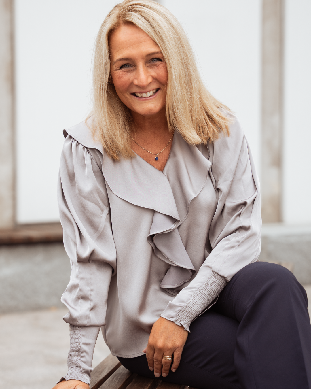 A middle-aged woman with blonde hair smiling, sitting on a wooden bench outdoors, wearing a light gray blouse and dark pants.