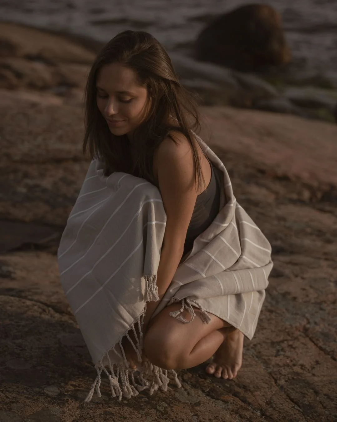 A woman with brown hair squatting on a rocky surface, wrapped in a beige striped blanket, wearing a black top, looking down with a gentle smile.