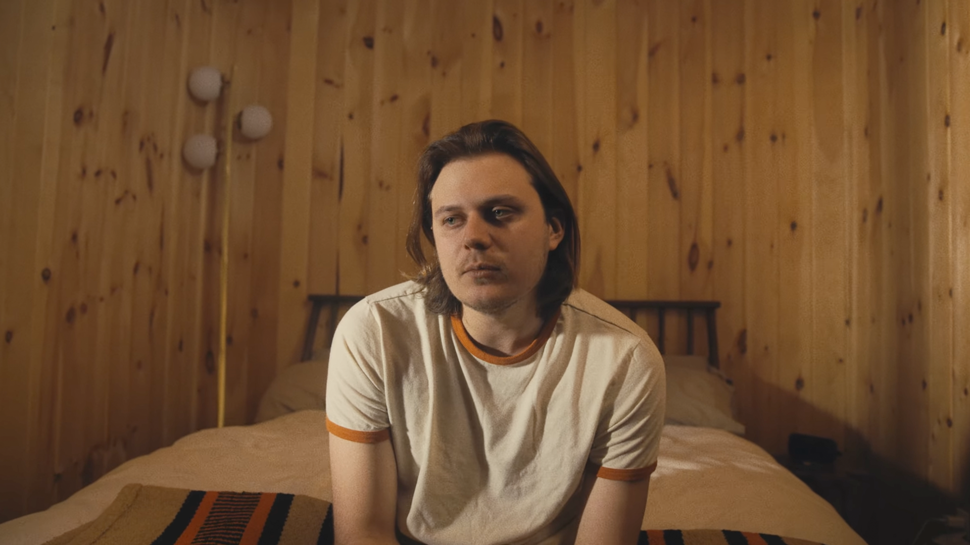 A man with shoulder-length brown hair and a serious expression, sitting on a bed in a room with wooden panel walls.