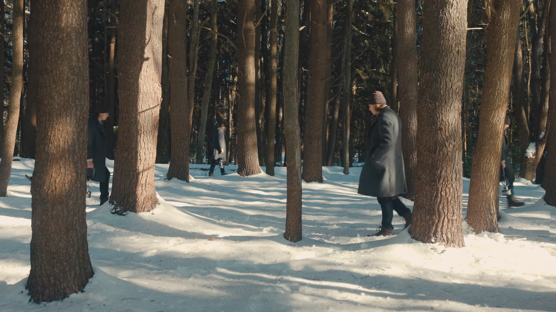 People wearing winter coats and hats walking through a snow-covered forest with tall trees.